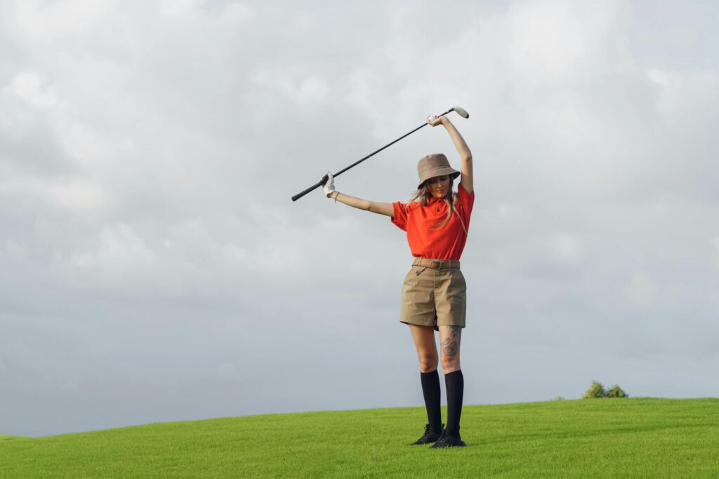 Woman golfer posing on a grassy course with a club and a stylish outfit under a cloudy sky.