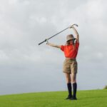 Woman golfer posing on a grassy course with a club and a stylish outfit under a cloudy sky.